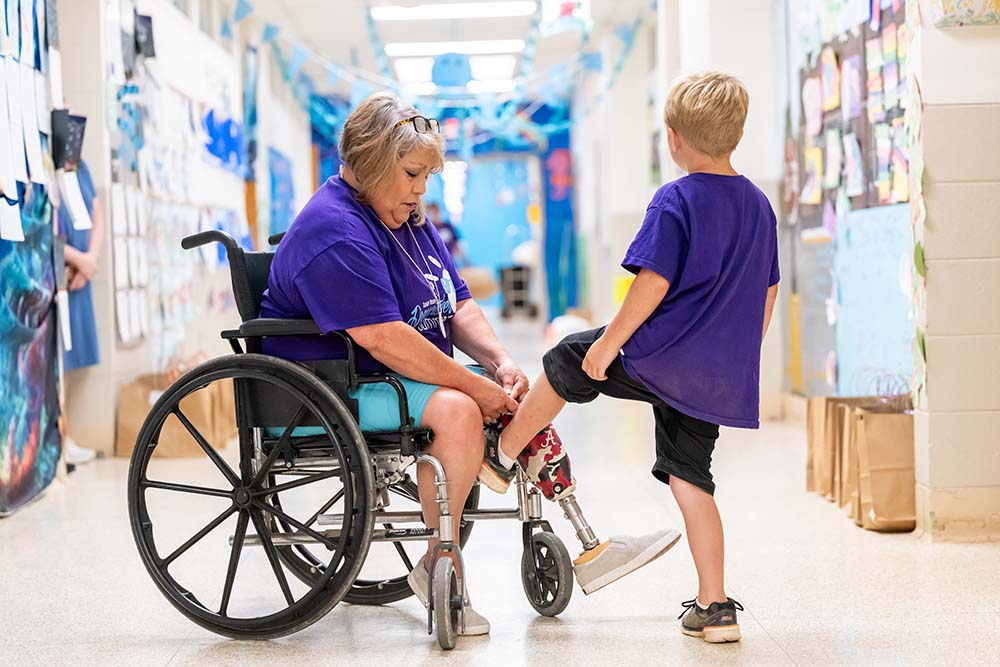 A woman with a prosthetic leg uses a wheelchair while helping a young boy tie his shoelace.