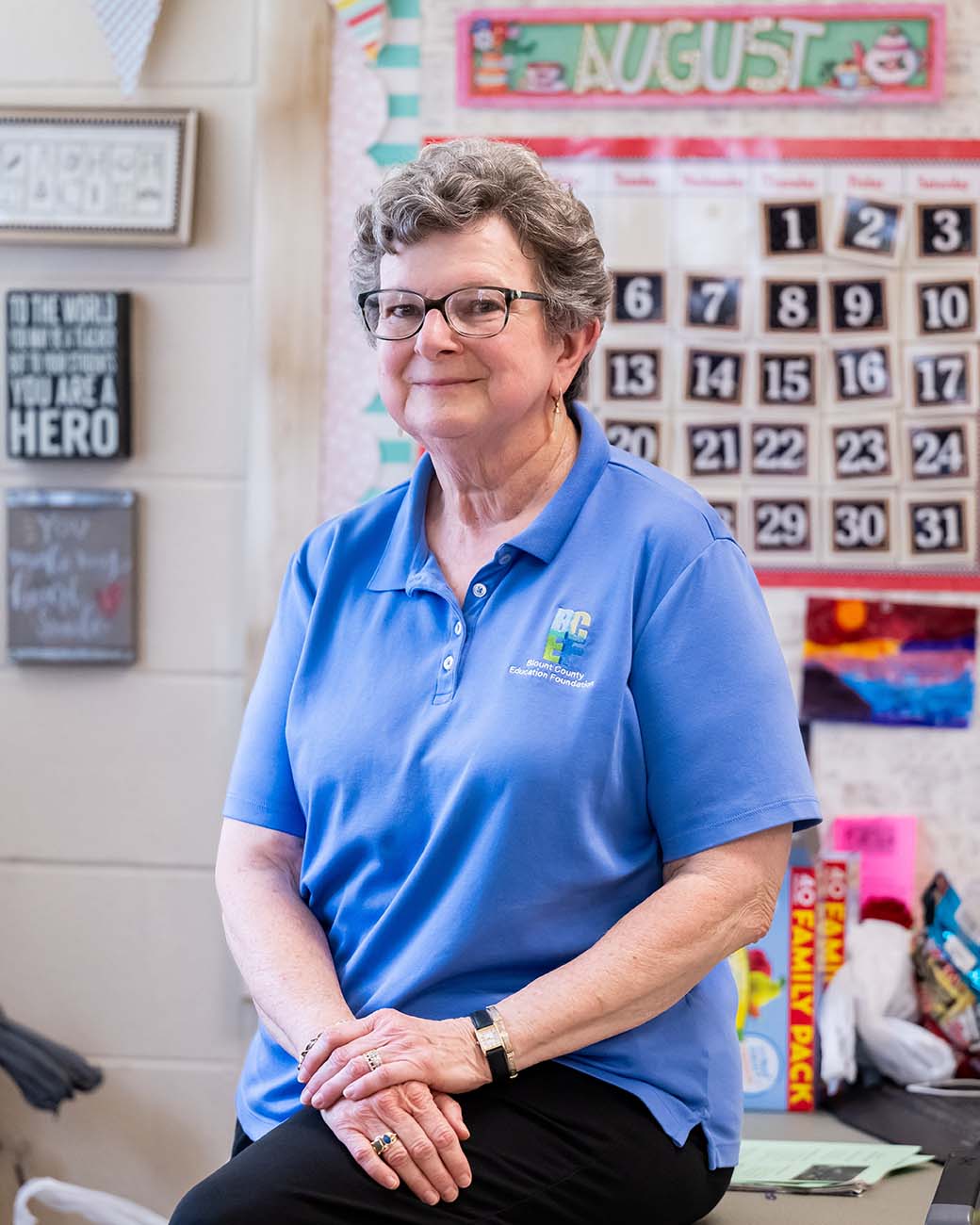 A woman with short gray hair wearing a blue polo shirt leans on a desk in a classroom.