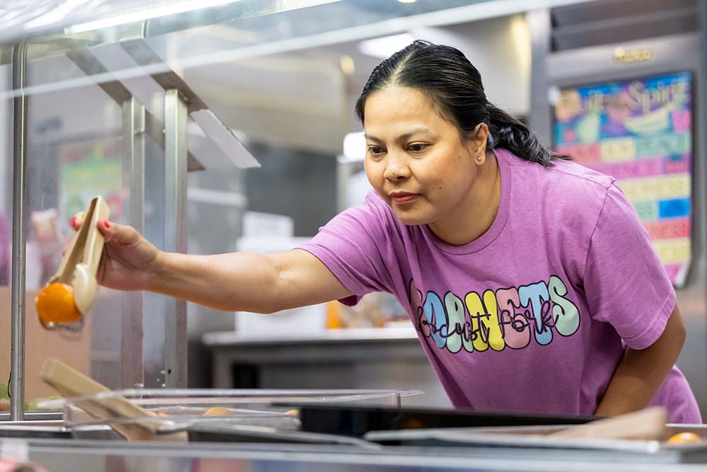 A woman in a pink t-shirt serves a tangerine from a cafeteria line.