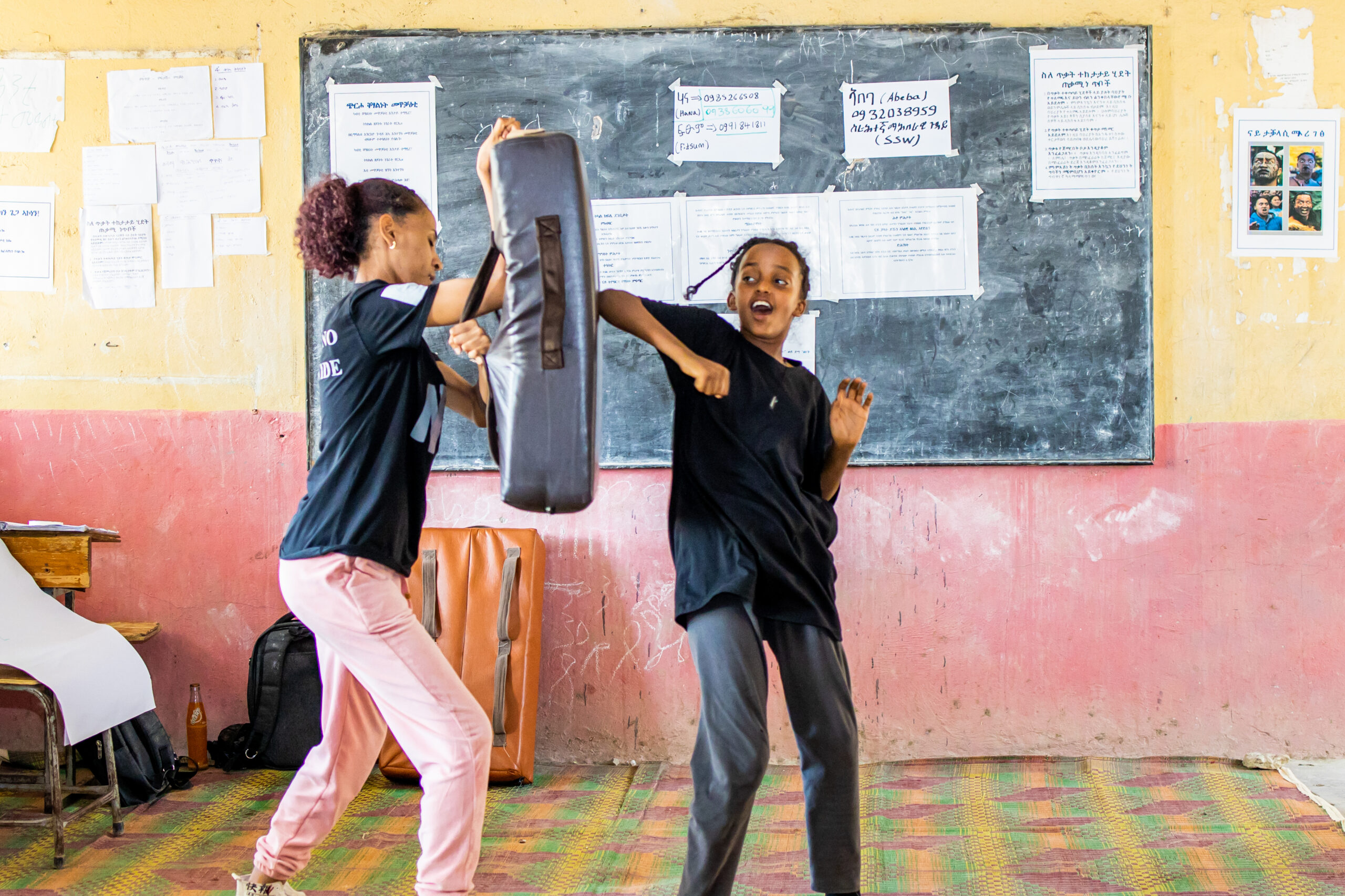 Young female student standing up hitting mat being held by trainer in middle of classroom in front of blackboard.