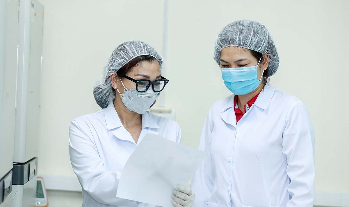 Two women wearing white lab coats, hair nets and medical face masks read a report.