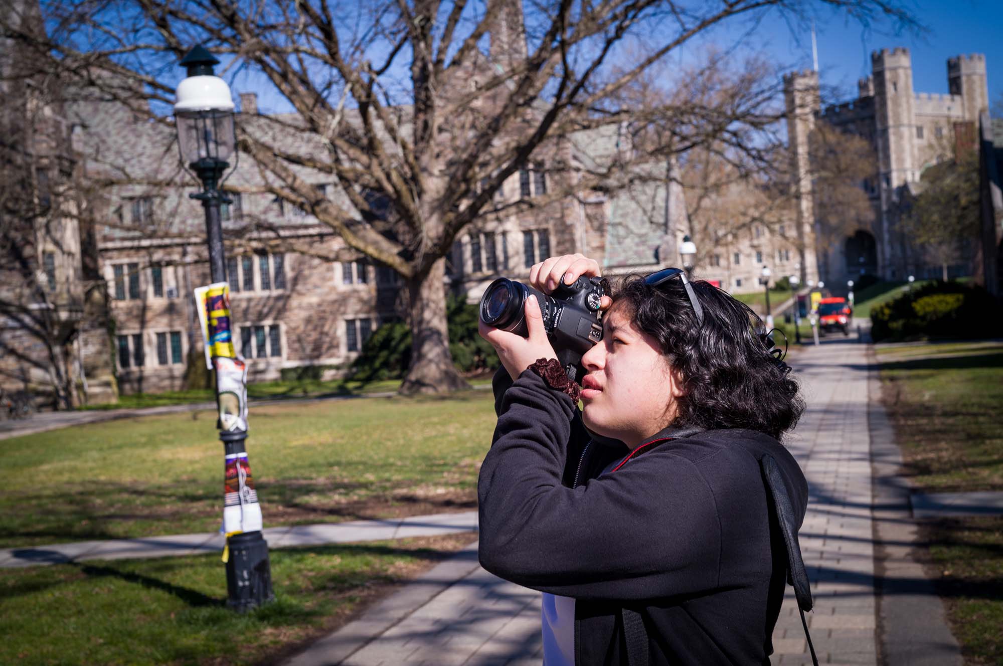 A teenager uses a DSLR camera to take a photo. She's standing in a courtyard surrounded by old academic buildings.