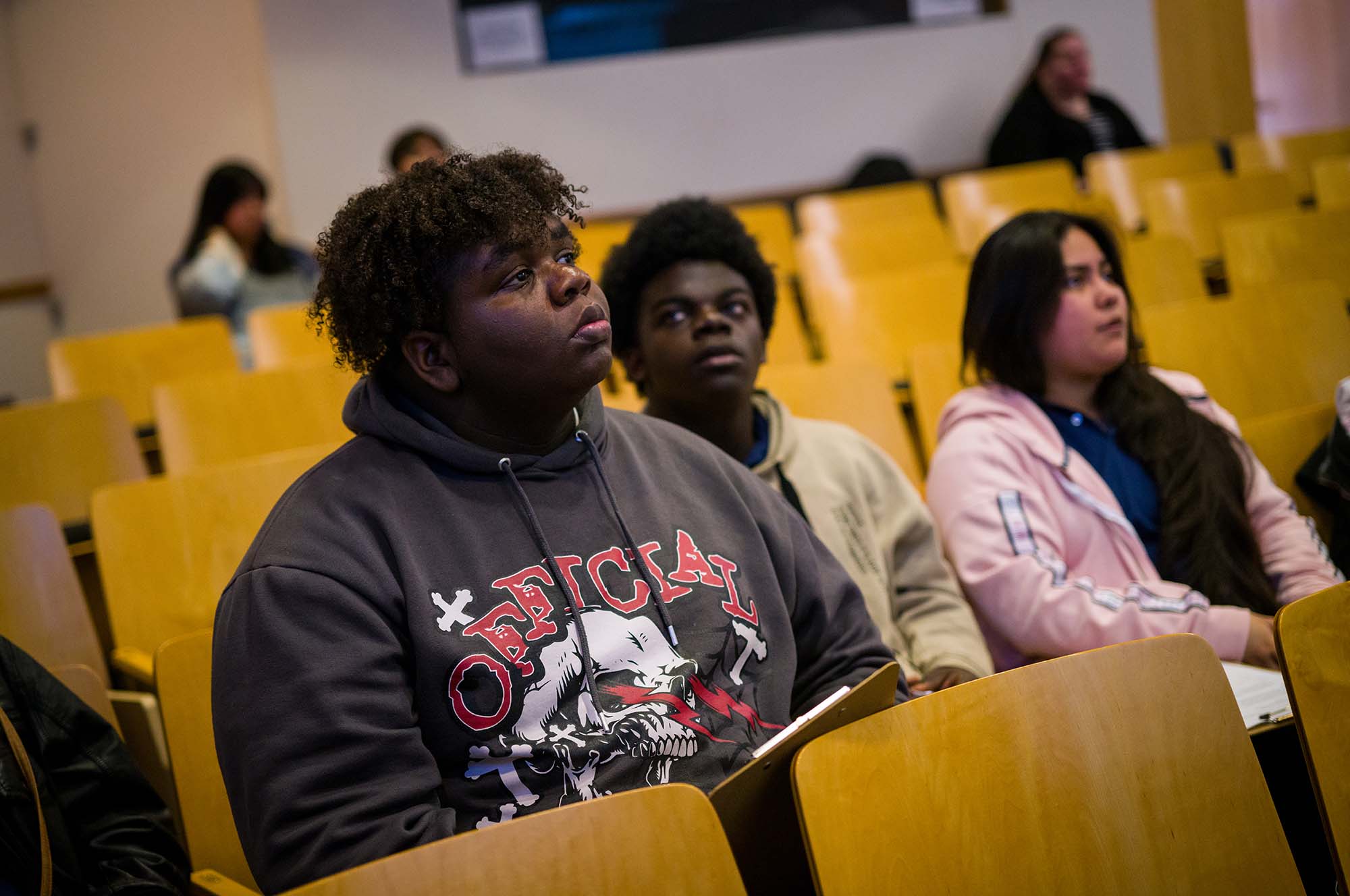 A student in a graphic sweatshirt sits in an auditorium seat and listens intently. More students are seating around him.