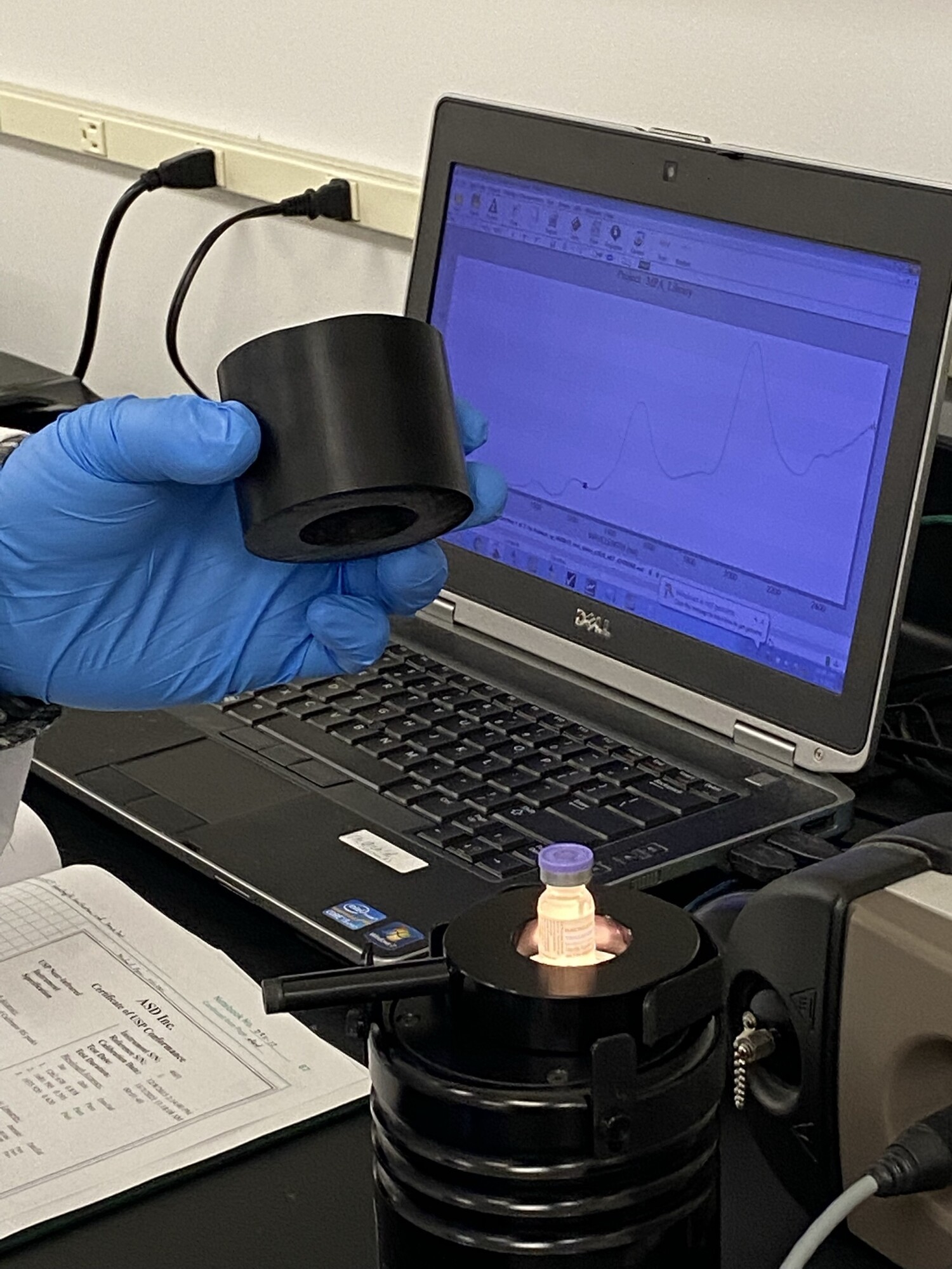 A lab device is held in front of a computer screen by a hand wearing a latex glove.