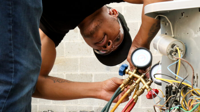 A young man in a black t-shirt and baseball cap works on a set of hoses attached to an HVAC unit.