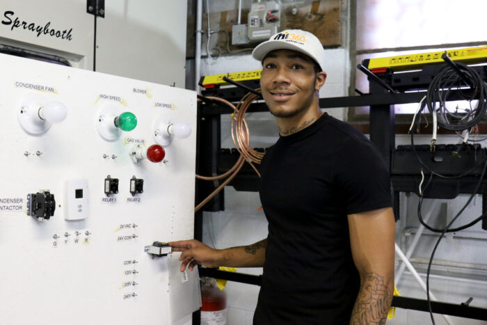 A young man in a black t-shirt and FHI 360-branded baseball cap flips a switch on a panel for a spray paint booth.
