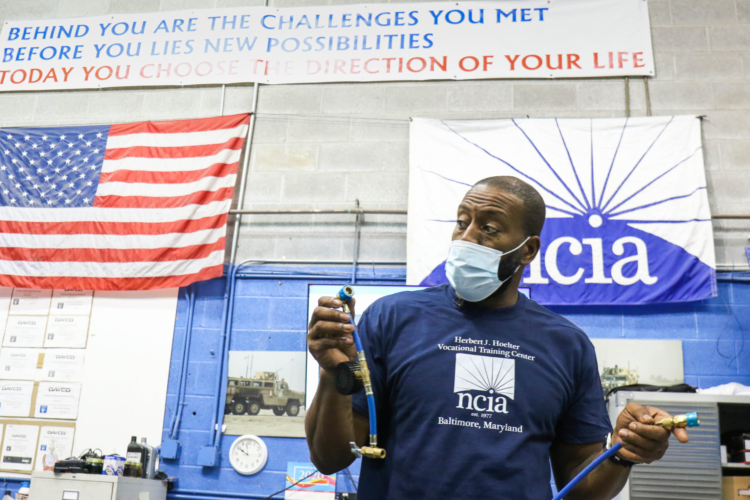 A man wearing a blue NCIA-branded t-shirt holds two mechanical hoses while instructing his class.