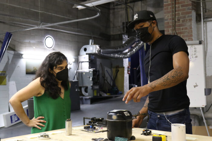 A young man wearing a black t-shirt speaks about tools sitting on a tabletop to a woman in a green tank top.