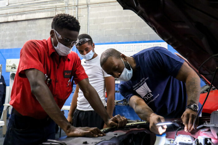 Two teenage students look into a car engine while the instructor points at something.
