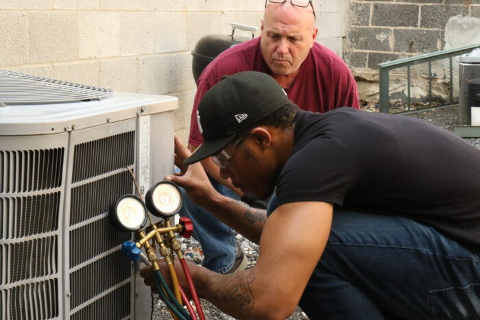 A young man in a black t-shirt and baseball cap works on a set of hoses attached to an HVAC unit while his teacher looks on.
