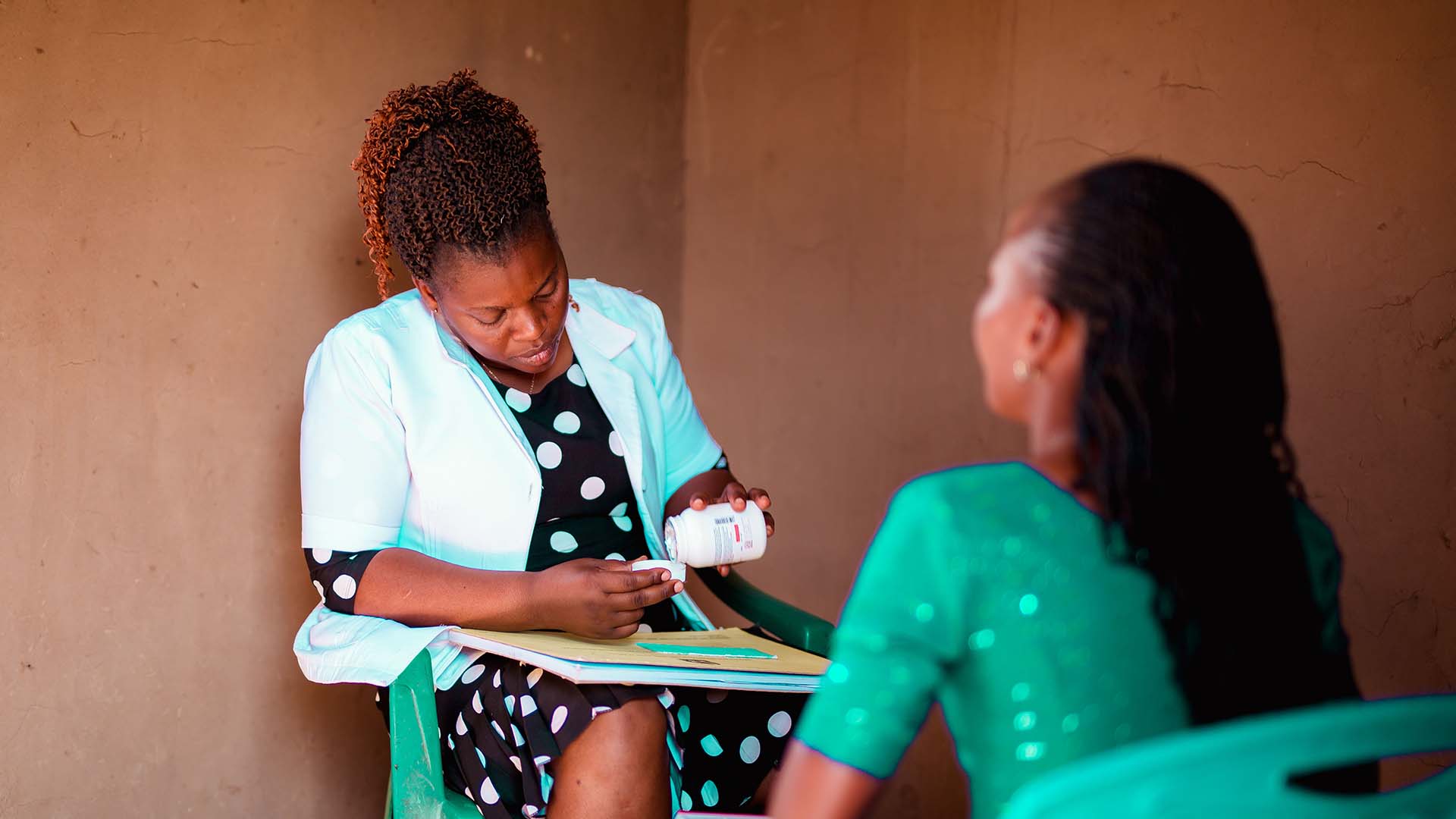 A woman wearing a white jacket dispenses medication from a bottle for another woman.