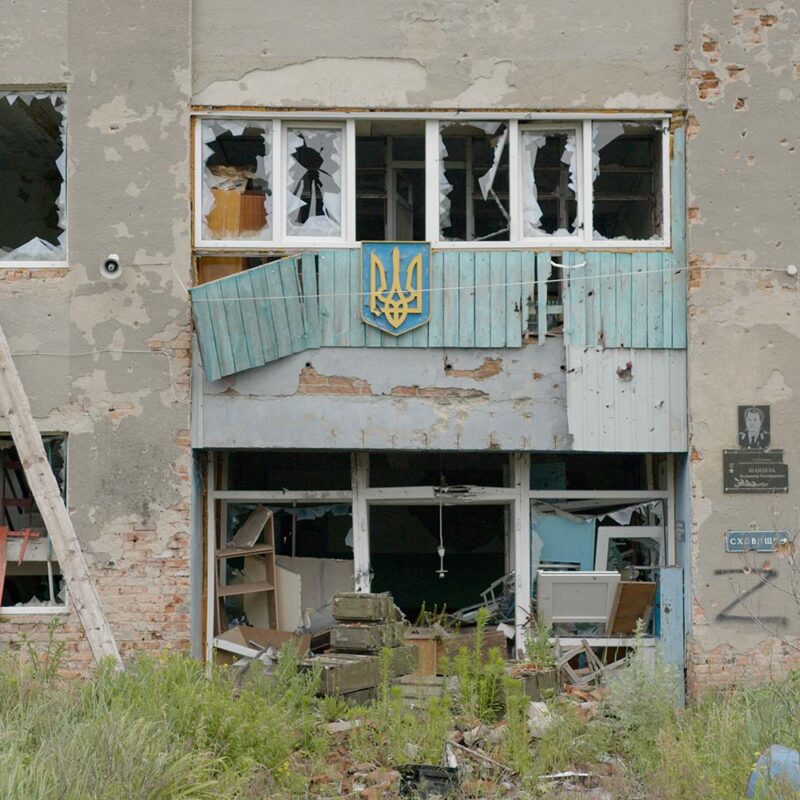 A gray concrete building with a blue and yellow Ukrainian emblem in the center, showing damage from bombs.