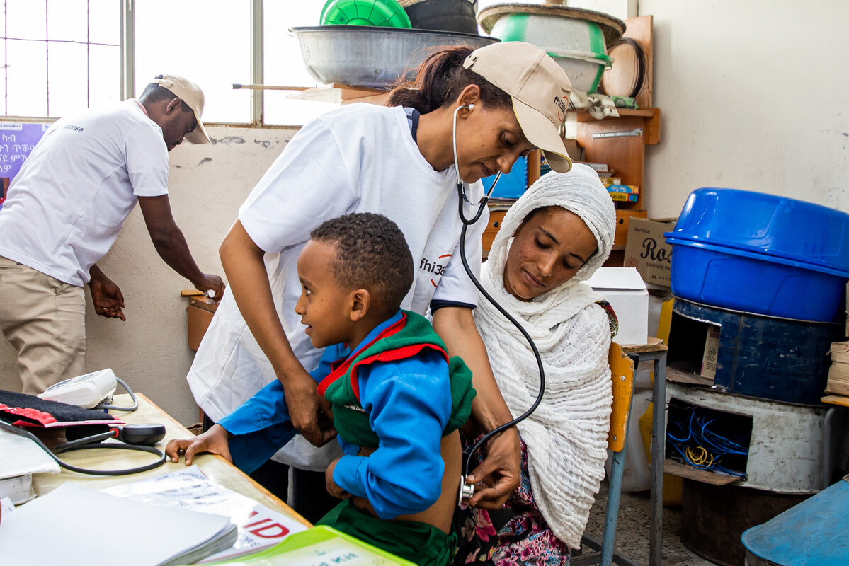 Mother holds her 6-year-old son as he is examined by health worker with stethoscope on his back.