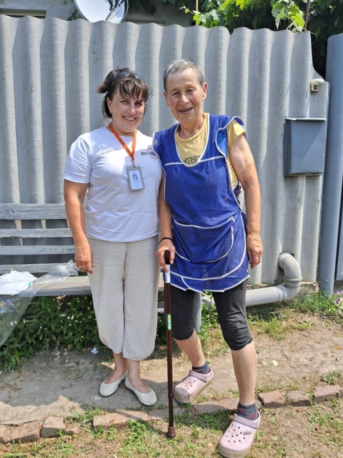 Two women are standing outside smiling, one is leaning on her cane, and the other is wearing her mobile medical team badge.