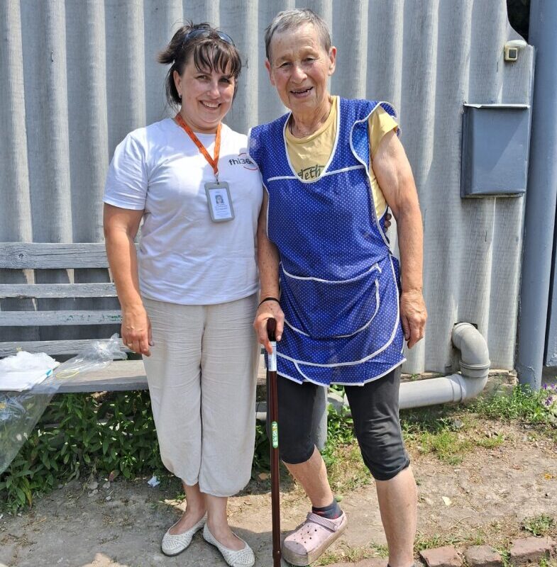Two women are standing outside smiling, one is leaning on her cane, and the other is wearing her mobile medical team badge.