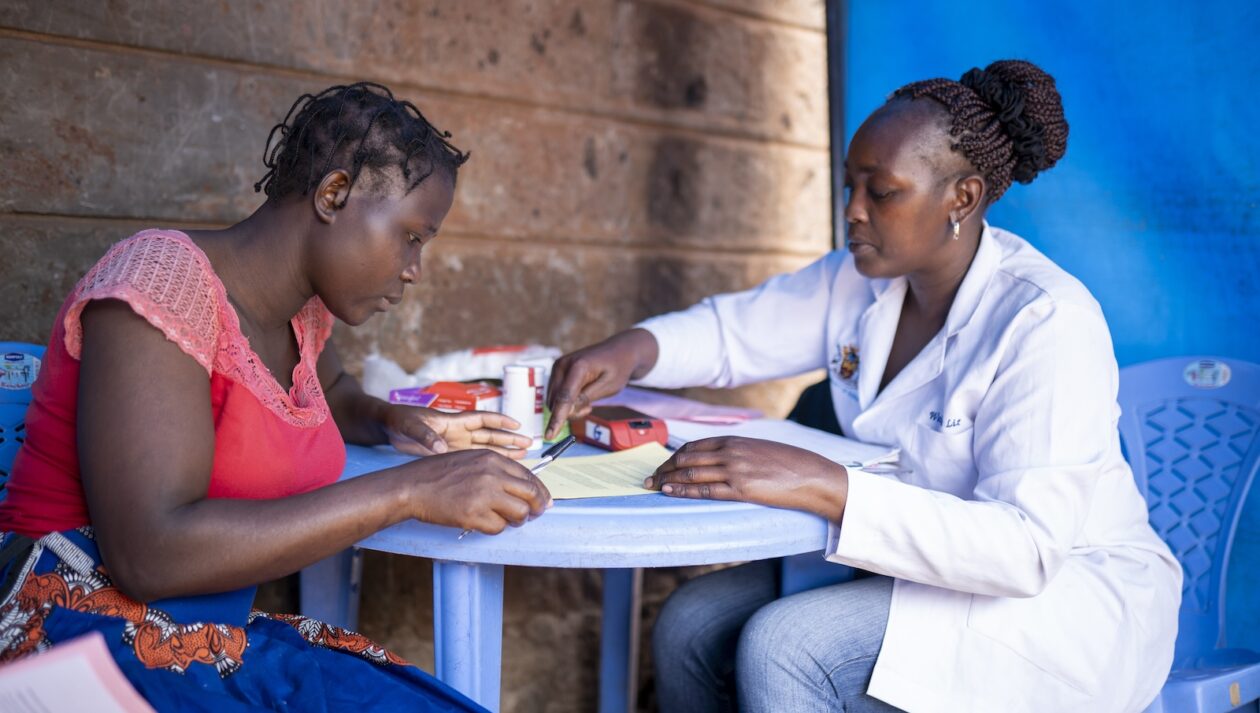 Two women, one in a lab coat, sit at a table together under a blue tent.