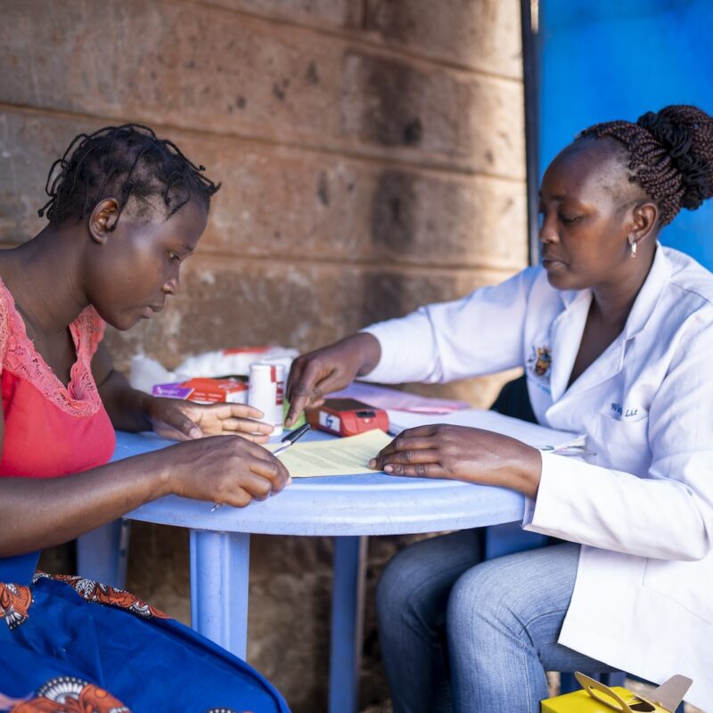Two women, one in a lab coat, sit at a table together under a blue tent.