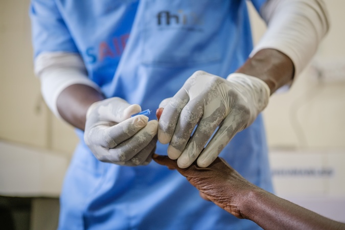 Two hands in medical gloves hold a lancet to collect blood from a man's hand.