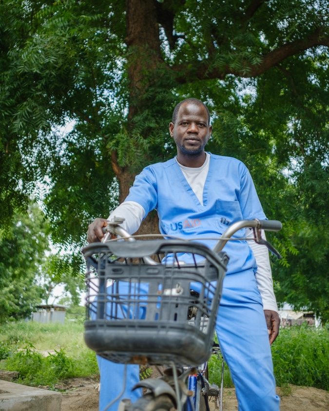 A man in scrubs leans on his bicycle in front of a tree.
