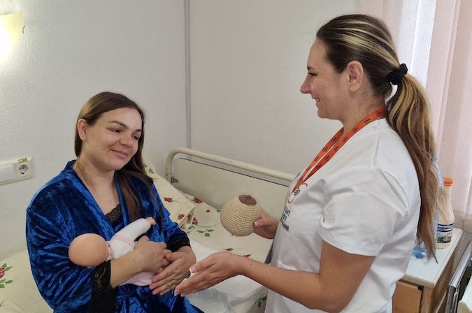 Yulia holds a crocheted breast and gestures to a sitting woman holding a babydoll.