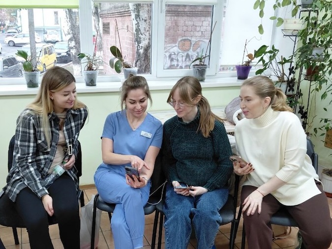 A row of women sitting in chairs look on as Mariia shows them the chatbot on her phone.