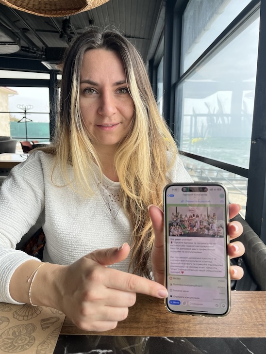 Yulia points to her phone screen, which displays the chatbot.