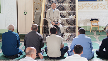 Imam Azalshoh speaks about TB to a group of seated men at Friday prayers.