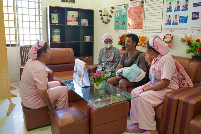 A dad holding his baby wrapped in blankets sitting in a waiting room with smiling nurses.
