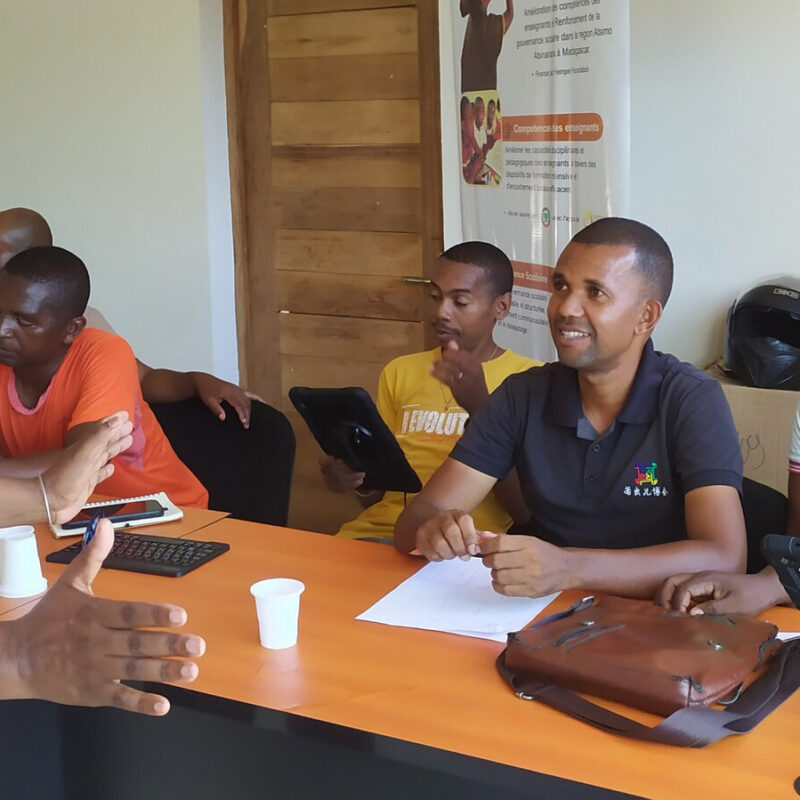 A teacher instructs a group students sitting at a large desk with various technology devices.