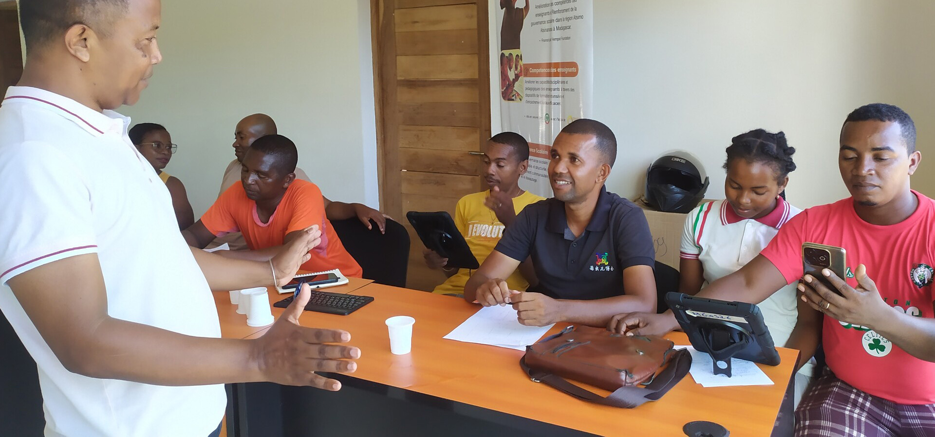 A teacher instructs a group students sitting at a large desk with various technology devices.