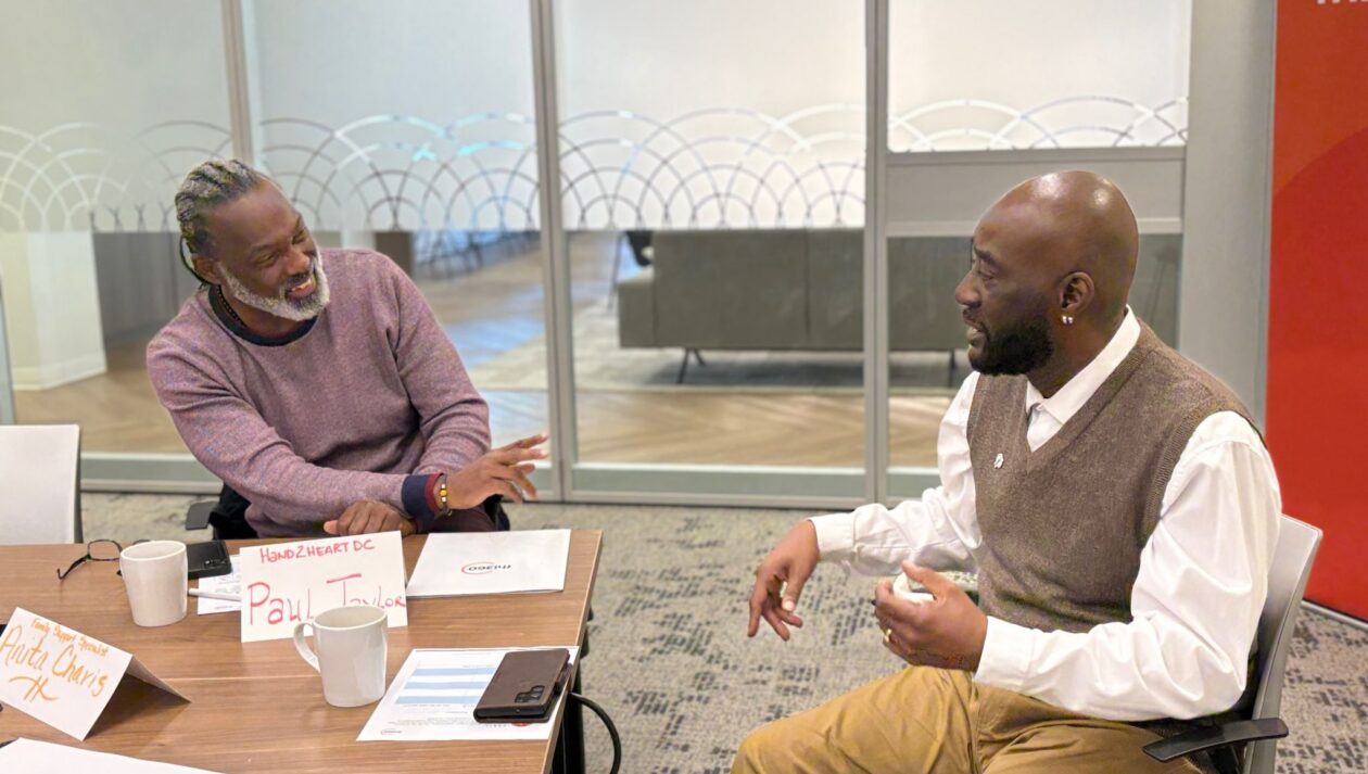Two men chat while sitting at a table in an office meeting room.