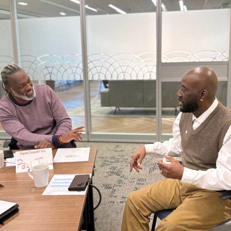 Two men chat while sitting at a table in an office meeting room.