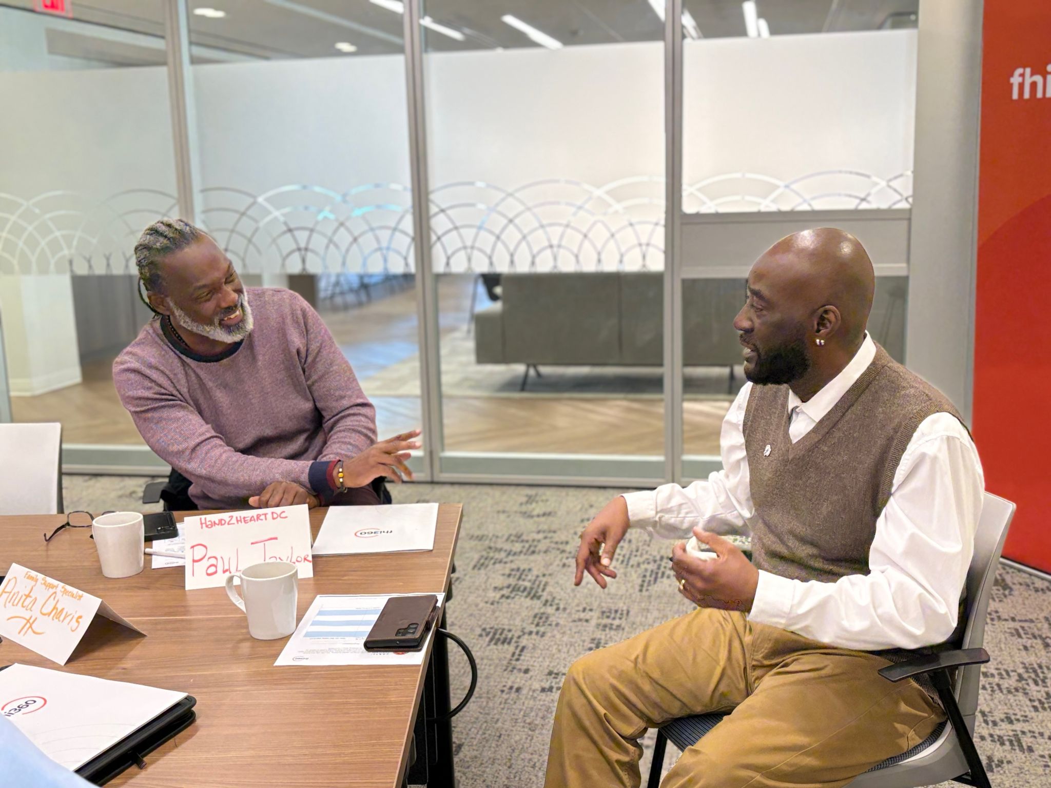 Two men chat while sitting at a table in an office meeting room.