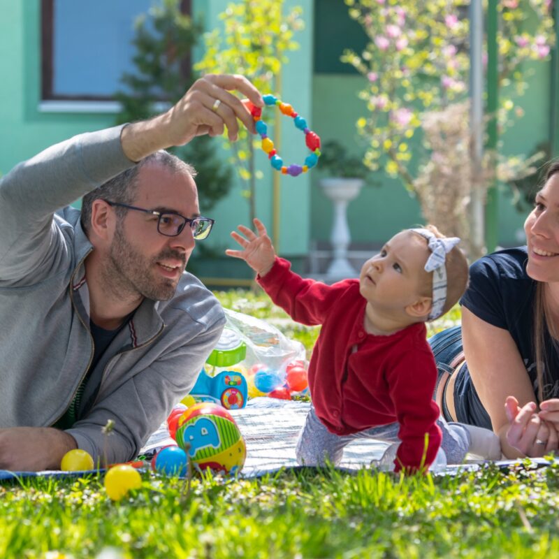 Parents in Serbia play with their child on a blanket outdoors in the sunshine as part of a playful parenting learning experience.