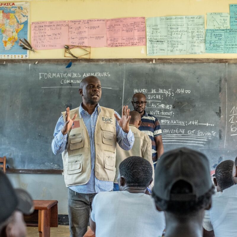A man, Marien, stands in front of a chalkboard instructing a classroom of people.