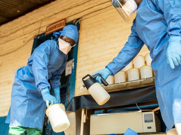 A technician in PPE gear sanitizes a container being held by another technician in PPE gear.